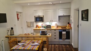 Een kleine keuken met witte kastjes, een magnetron, een broodrooster en een spoelbak. Er staat een houten tafel met een geruit tafelkleed en twee stoelen.