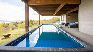 A rectangular swimming pool with a wooden pergola overhead, a grassy area with a fire pit, and a view of mountains in the background.