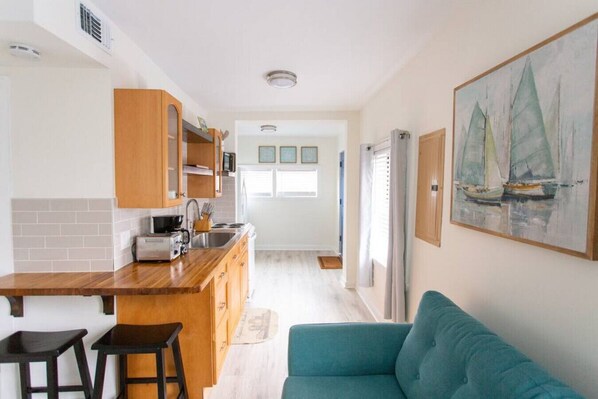 A compact kitchen with wooden cabinets, a built-in microwave, and a sink. There is a teal sofa and a painting of sailboats on the wall.