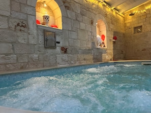 Un área interior con piscina de paredes de piedra, un jacuzzi, nichos abovedados e iluminación con luces colgantes.