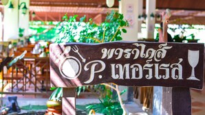 Une enseigne en bois, ornée d’écritures thaïlandaises et de symboles représentant une fourchette, un couteau et un verre à vin, indiquant un espace réservé aux repas.