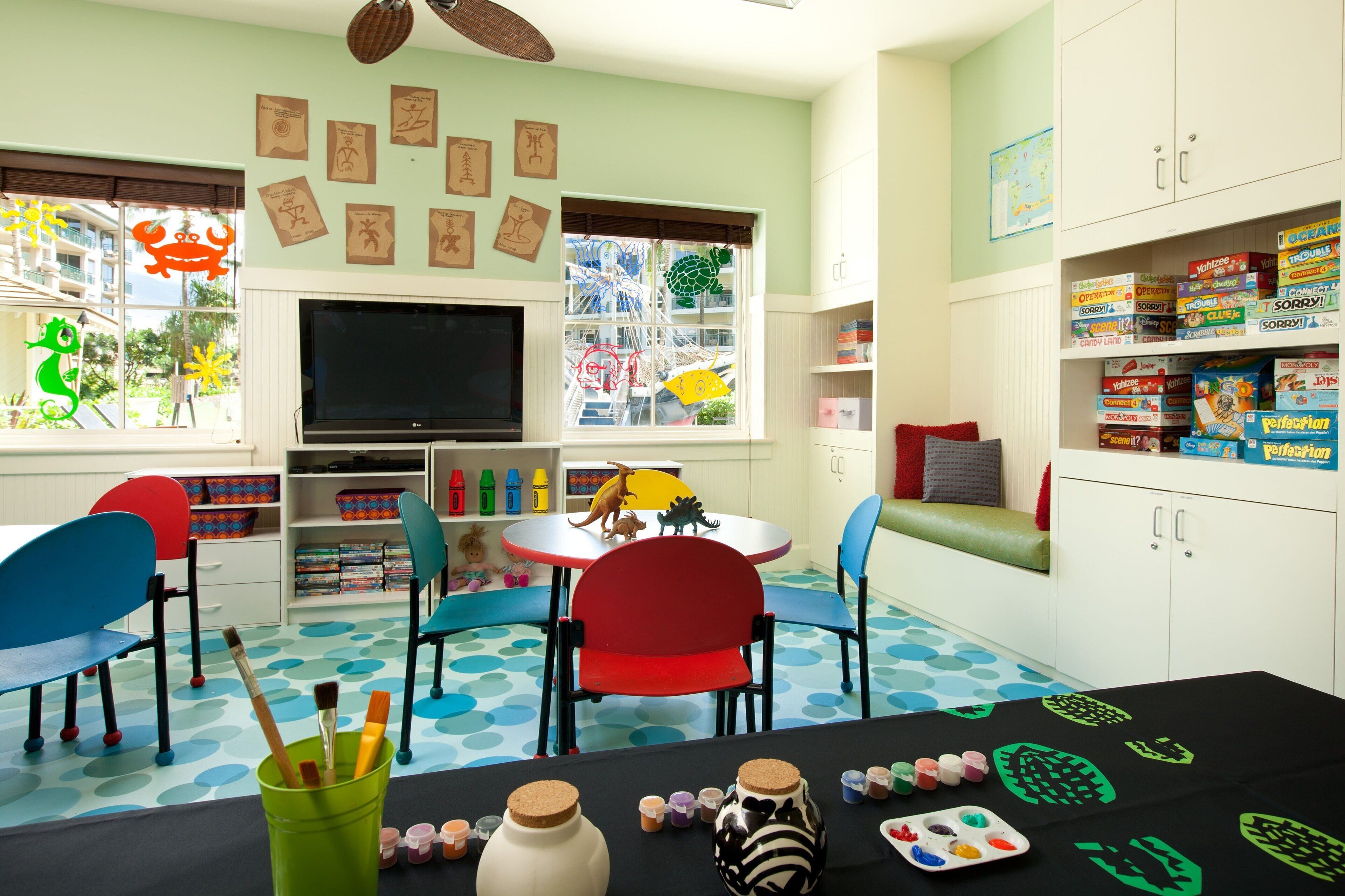 Una sala de juegos para niños con televisor, sillas coloridas, una mesa con materiales de arte y una ventana con vista a un edificio.