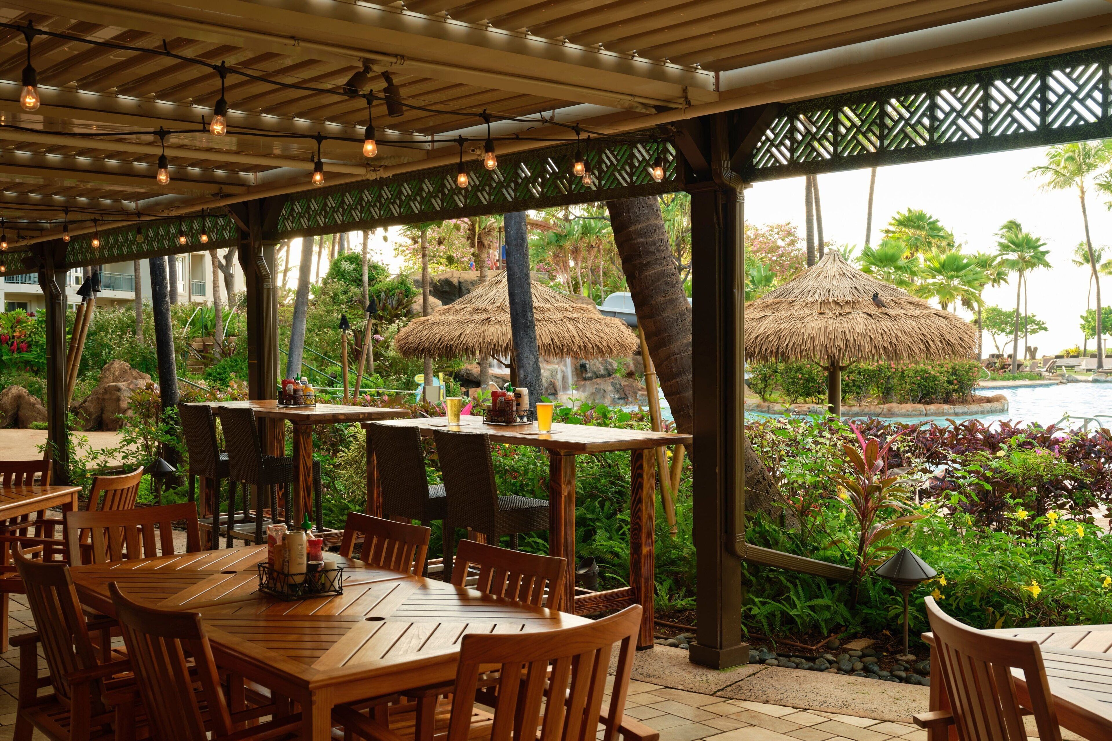 Un espace de restauration en plein air avec des tables et des chaises en bois, des parasols en chaume et une vue sur une piscine et des palmiers.