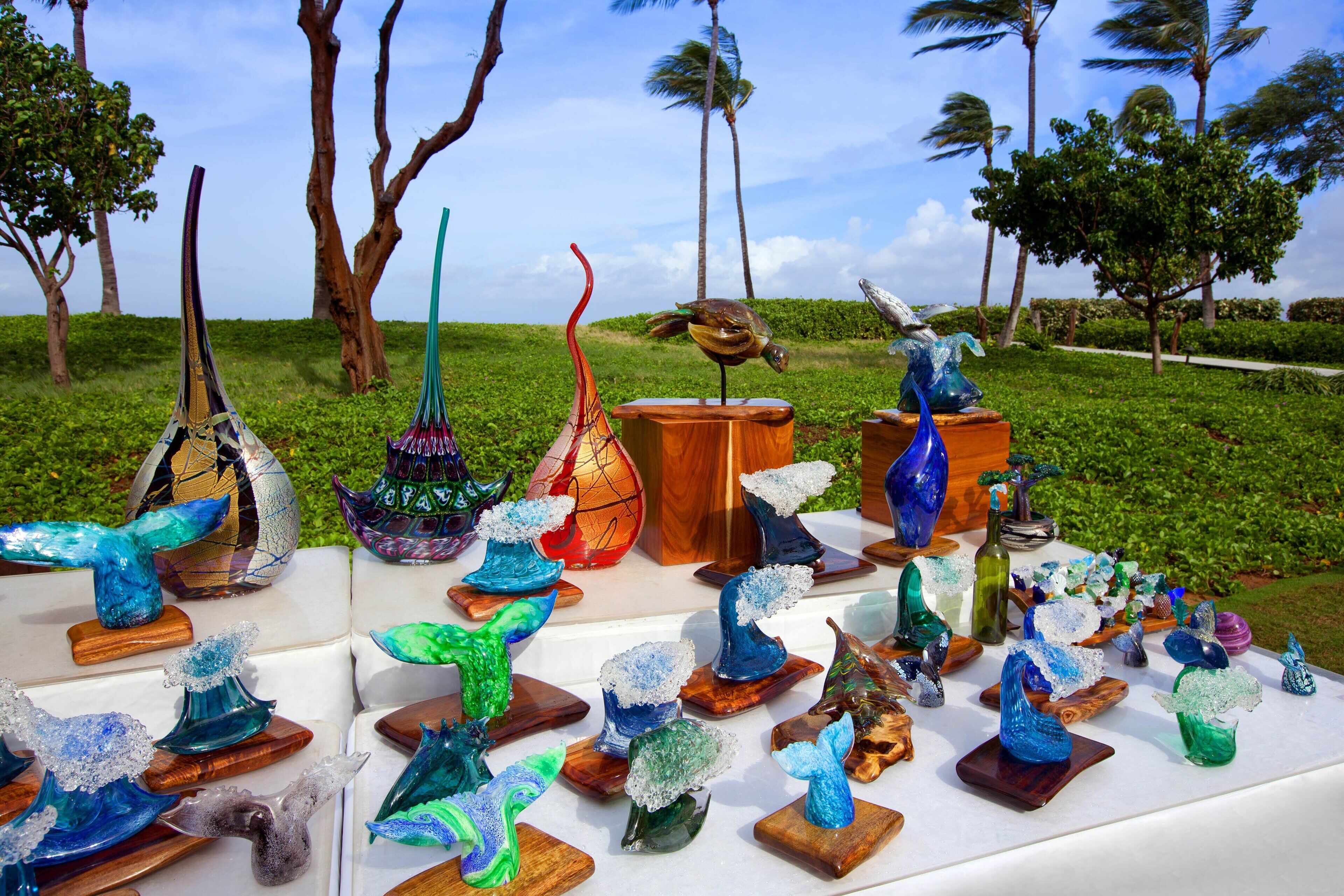 A display of glass art pieces on wooden stands in an outdoor setting with trees and a clear sky in the background.