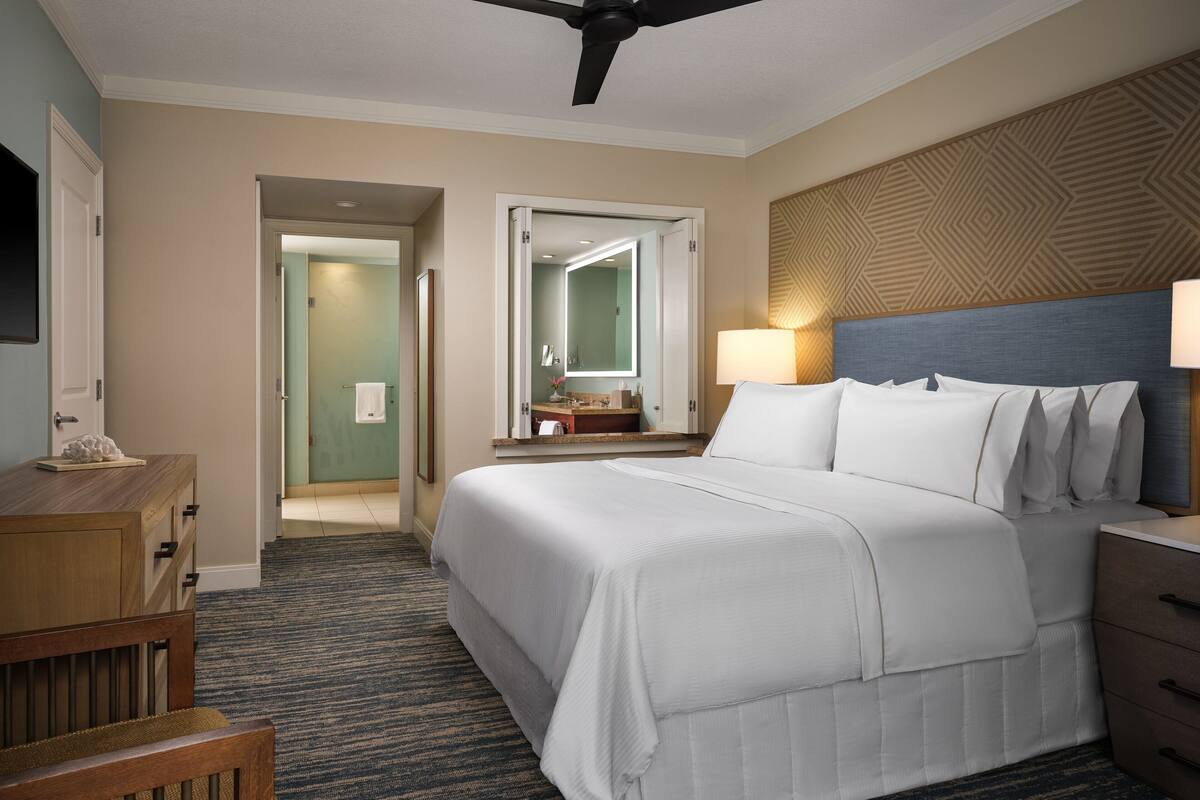 Une chambre d’hôtel avec un grand lit, un bureau, une chaise, un miroir et un ventilateur de plafond.
