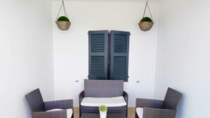 A small outdoor seating area with wicker furniture and a table, two hanging baskets with plants, and a pair of blue shutters.