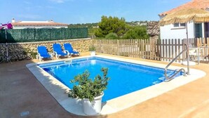 A swimming pool with blue water, surrounded by a stone wall and a wooden fence. There are blue lounge chairs and a potted plant near the pool.