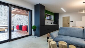 A modern living room with a grey sofa, wooden stumps, a kitchen with black cabinets, and a green accent wall.