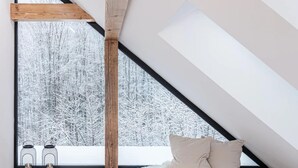 A high-ceilinged room with a large window overlooking a snowy forest. A wooden beam supports the ceiling, and a white fur rug is on the floor.