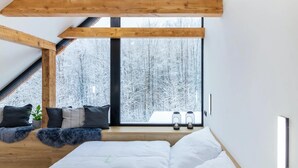 A modern bedroom with a sloped ceiling, wooden beams, a large window with a snowy view, a bed with white bedding, and two wall-mounted lanterns.