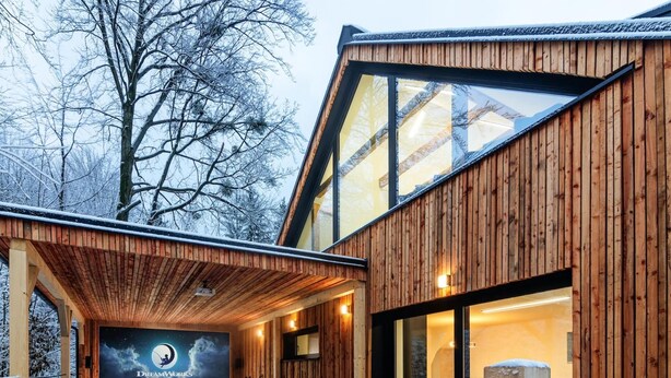 A wooden cabin with a covered outdoor seating area, a large window, and a view of the surrounding trees.
