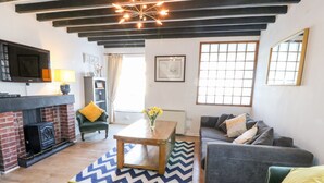 A living room with a fireplace, a sofa, a table, and a window with natural light. - Malltraeth Cottage (Bodorgan)