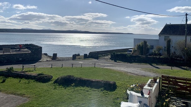 A coastal view with a stone structure, a grassy area, and a building with a tiled roof. - Malltraeth Cottage (Bodorgan)