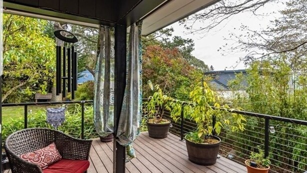 Una terraza de madera con plantas en macetas, una silla de mimbre con cojín rojo y una barandilla de metal.