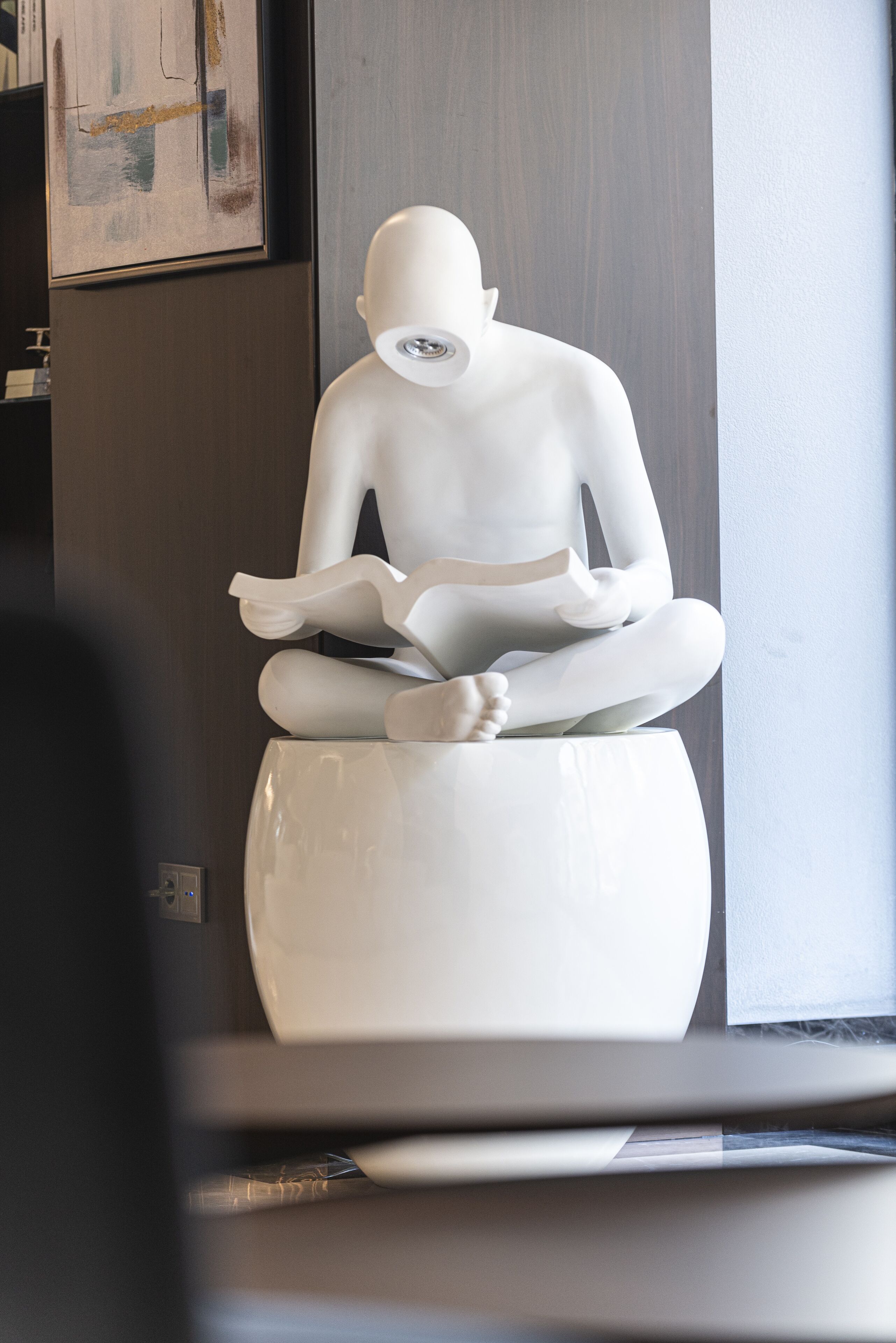 Une statue blanche représentant une figure en méditation, placée sur un piédestal dans une pièce dont un mur est recouvert de lambris sombres et où est accrochée une peinture abstraite encadrée.