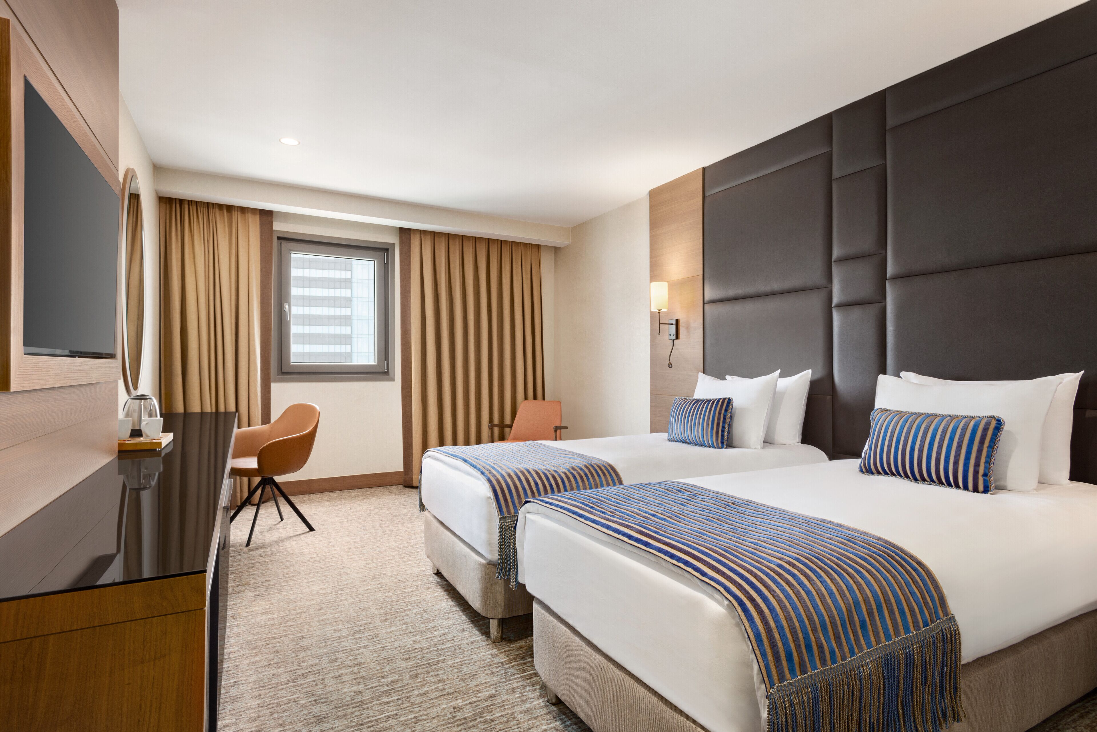 A hotel room with two beds, a desk, a chair, and a large window with curtains.