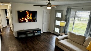 Una sala de estar con televisor de pantalla plana, piso de madera oscura, ventilador de techo y un ventanal con vistas al exterior.