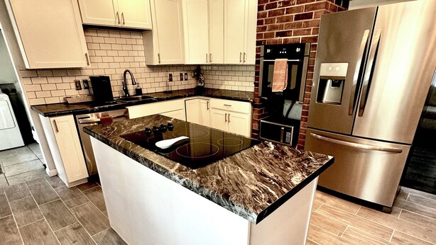 Una cocina moderna con armarios blancos, encimera de granito, refrigerador de acero inoxidable y pared decorativa de ladrillo.