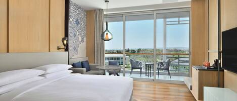 Una habitación de hotel moderna con una cama grande, un balcón con vistas, una zona de comedor y un televisor.