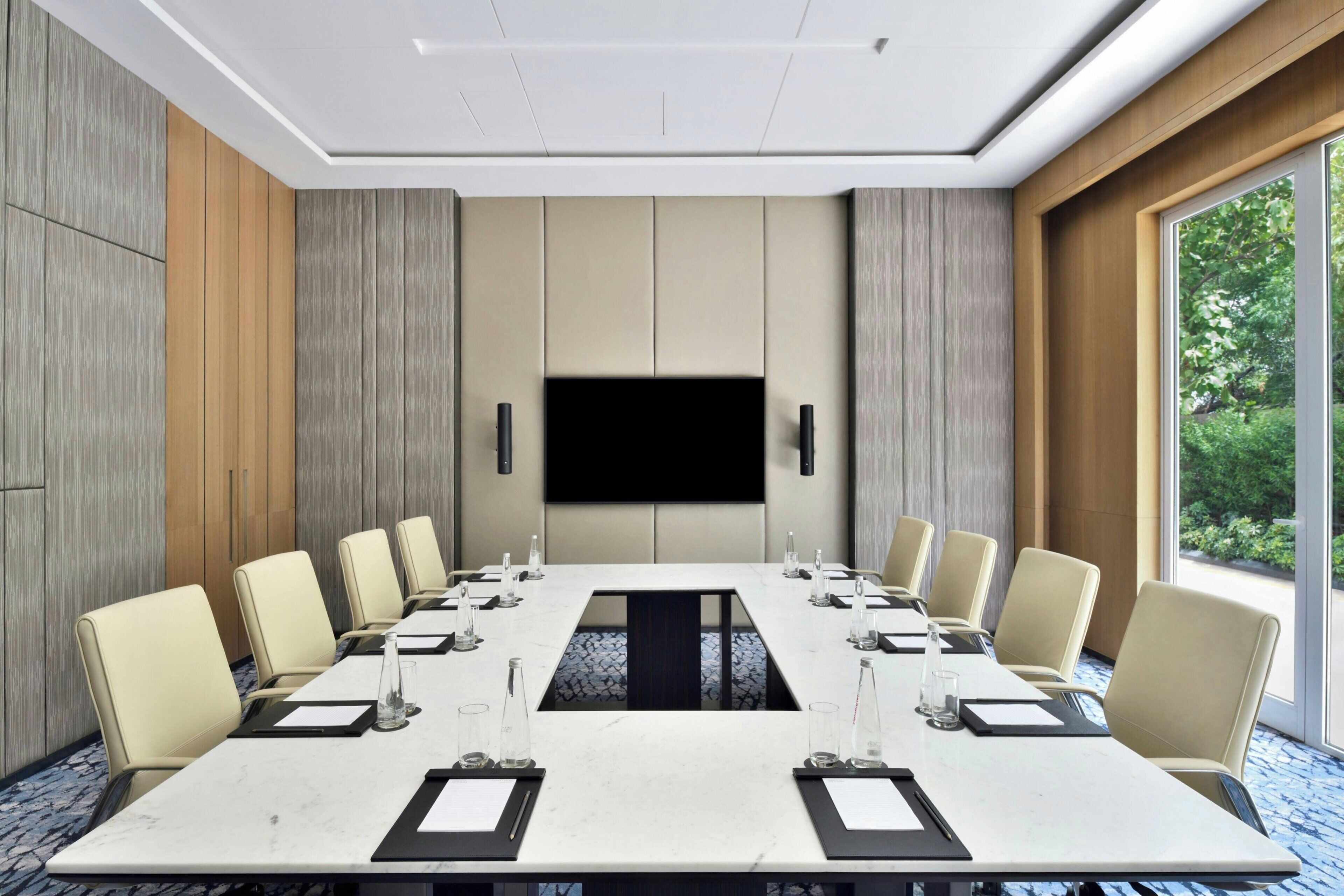 A conference room with a long table, chairs, a flat-screen TV, and a large window with a view of greenery.