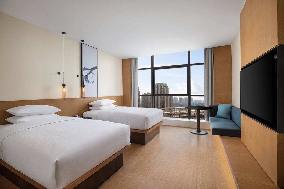 Ein Hotelzimmer mit zwei Betten, einem Sofa, einem Fernseher und einem großen Fenster mit Blick auf die Stadt.