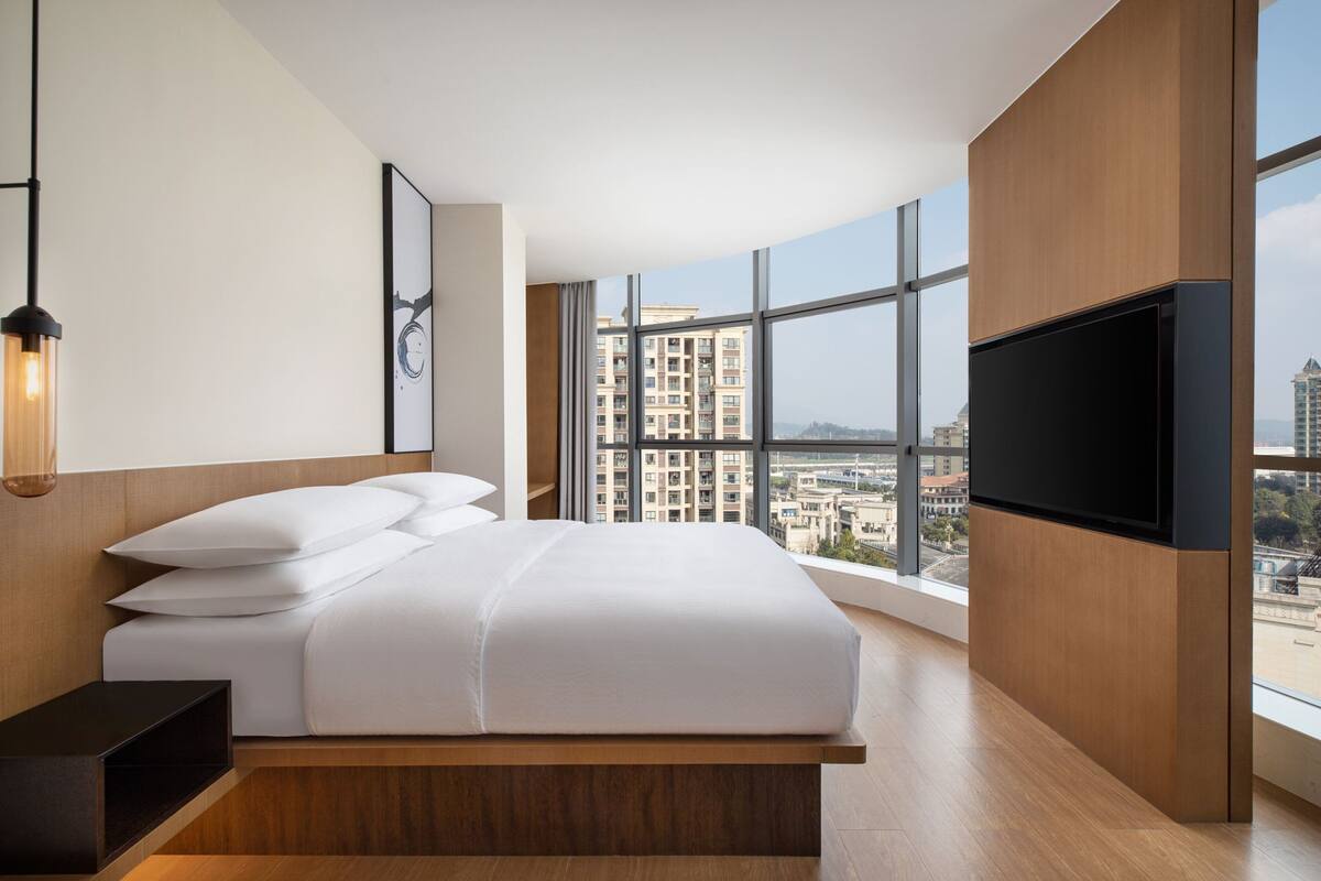 Ein modernes Hotelzimmer mit einem großen Bett, einem Flachbildfernseher und Blick auf die Stadt durch große Fenster.