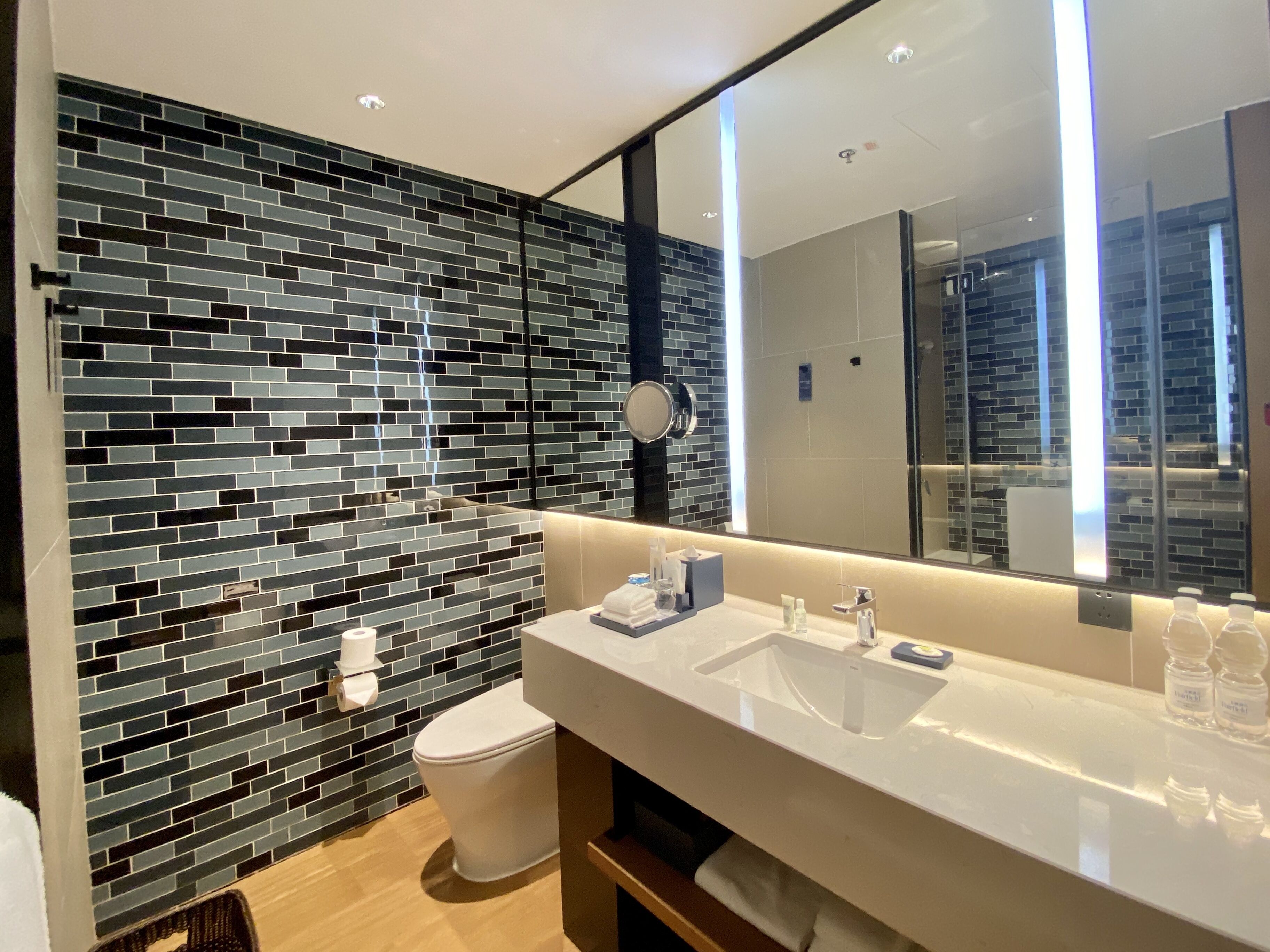 Un baño moderno con pared de mosaicos negros y grises, un inodoro blanco, un espejo grande y un lavamanos blanco con artículos de tocador.