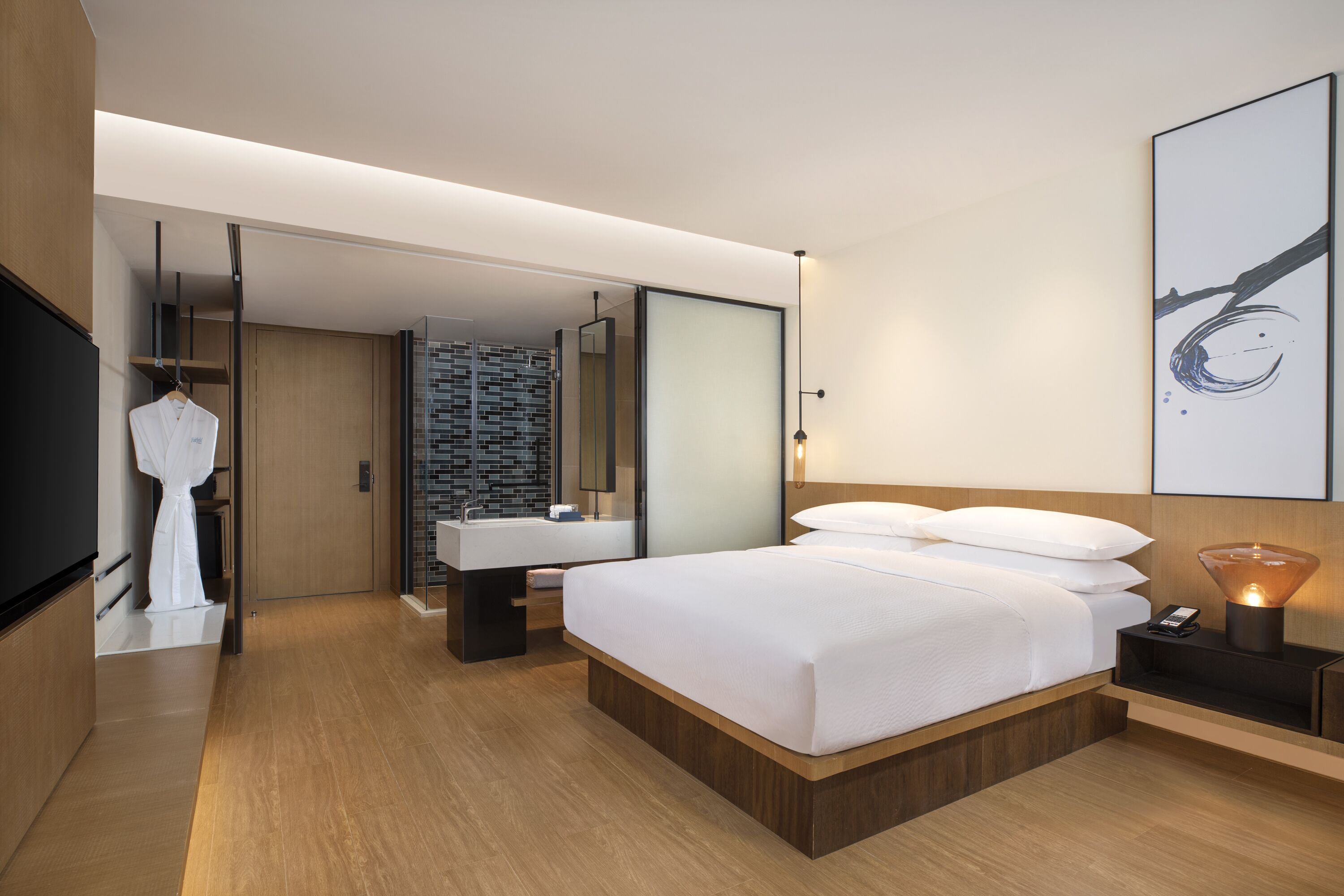 A modern hotel room with a large bed, a bedside table with a lamp, a full-length mirror, and a bathroom with a sink and toilet.