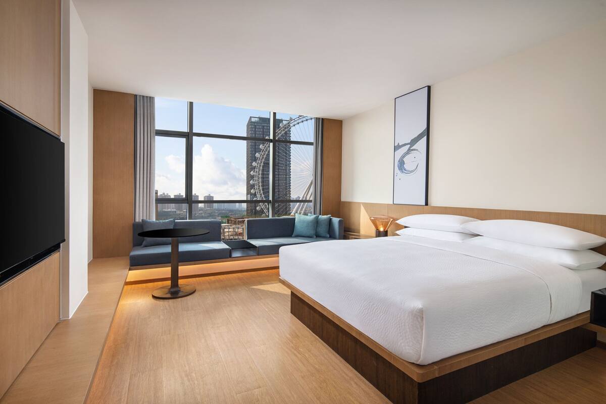 Ein modernes Hotelzimmer mit einem großen Bett, einer Sitzecke mit Sofa und Sesseln, einem Fernseher und einem großen Fenster mit Blick auf die Stadt.