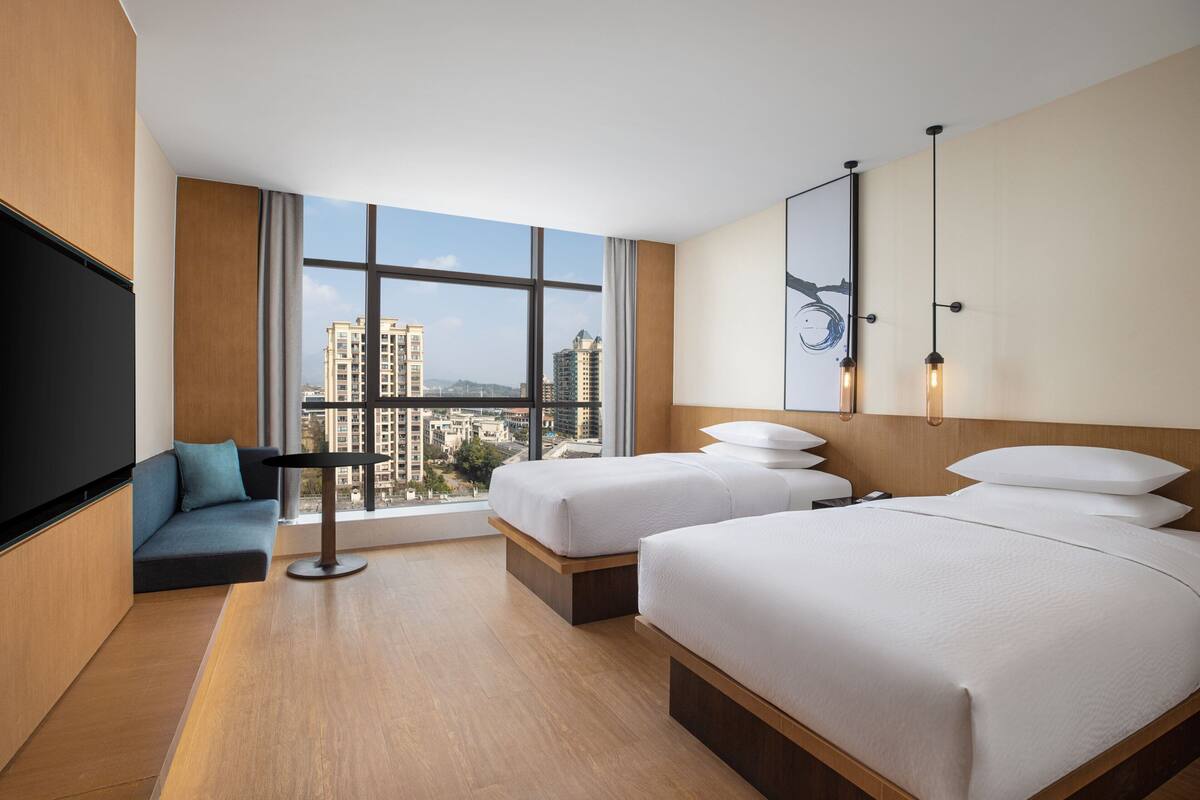 Ein Hotelzimmer mit zwei Betten, einem Schlafsofa, einem Fernseher und einem großen Fenster mit Blick auf die Stadt.