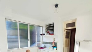 A compact kitchen with a dining area, a refrigerator, and a window with blinds. - Airy studio apartment in Copacabana, 4 blocks from the beach. (Rio de Janeiro)