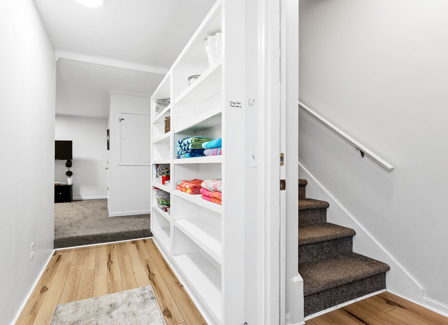 A hallway with a white door leading to a room, a staircase, and a storage unit with folded blankets.