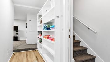 A hallway with a white door leading to a room, a staircase, and a storage unit with folded blankets.