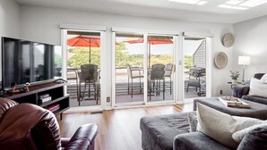 A living room with a TV, leather couch, and a sliding glass door leading to an outdoor patio with chairs and umbrellas. - Compass Pointe Condominium - Steps to downtown, close to Lake Michigan (Grand Haven)