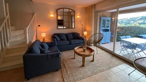 A living room with a dark blue sofa, a small round table with a candle, and a chair. There is a staircase on the left and a view of a blue cabinet and outdoor furniture through a glass door. - 🏖️ Villa - Swimming pool - 6 people - Near beaches & golf - Wifi (Sainte-Maxime)