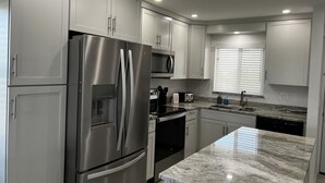Una cocina moderna con electrodomésticos de acero inoxidable, armarios blancos, encimera de granito y una ventana con persianas.