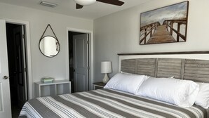 Una cama bien hecha con ropa de cama a rayas, un cabecero de madera, una lámpara de noche y un cuadro de un paseo marítimo de playa en la pared.