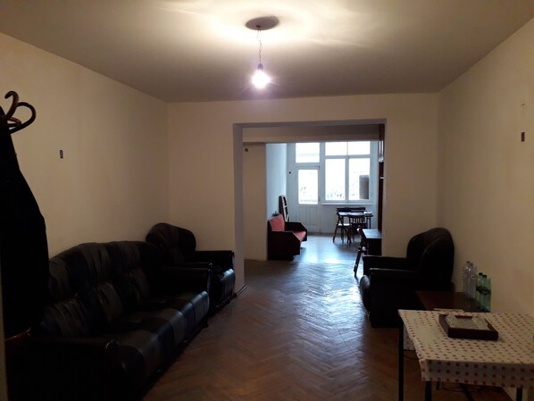 A living room with a black leather sofa, a small table with a white tablecloth, and a wooden floor. There is a window with a view of the outside.