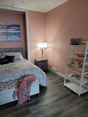 Een slaapkamer met een bed, nachtkastje, lamp, plank met handdoeken en een kleurrijk dekbed.