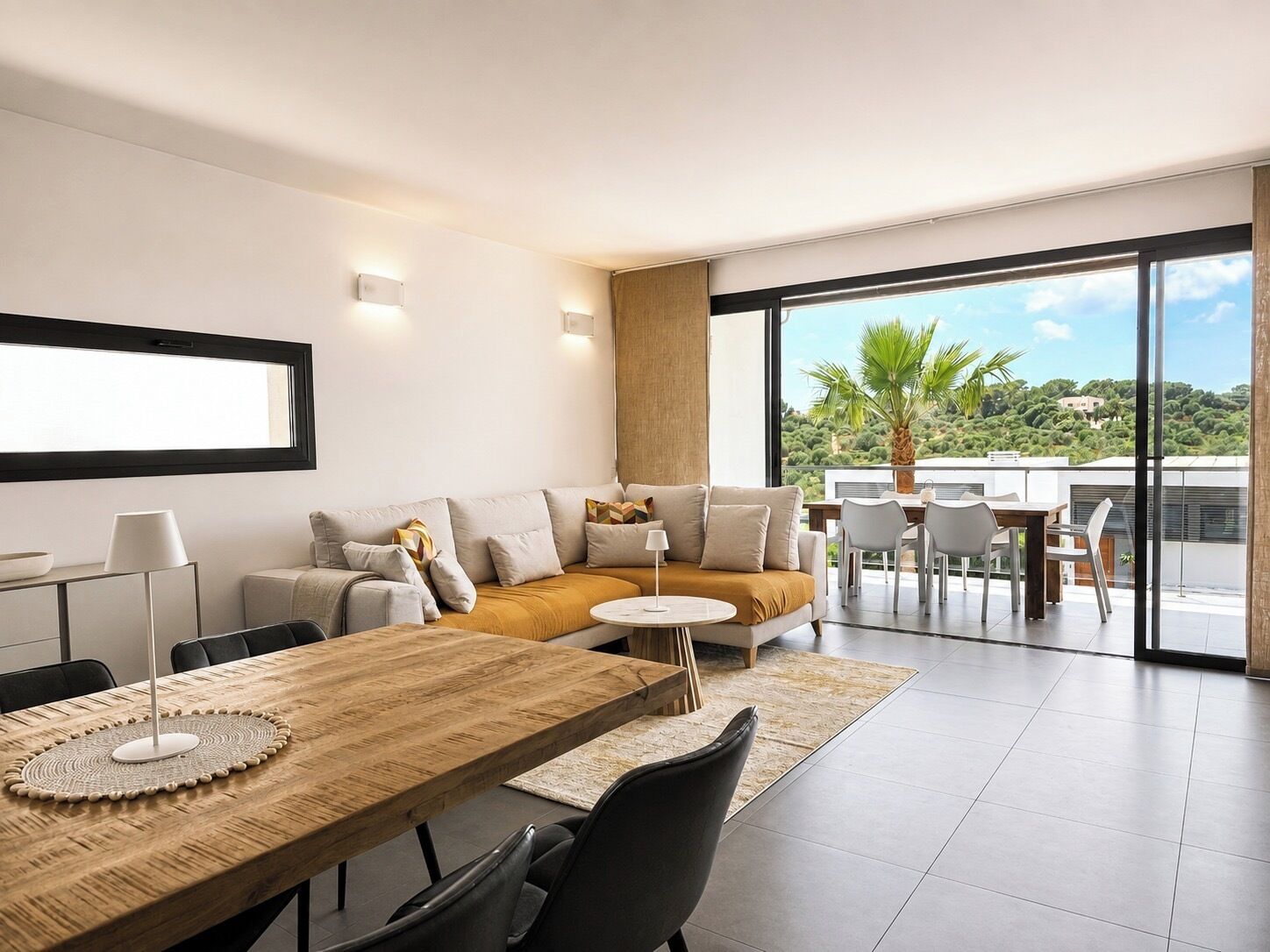 Un salon moderne comprenant un canapé, une table basse et un coin repas. Une grande fenêtre offre une vue sur l’extérieur.