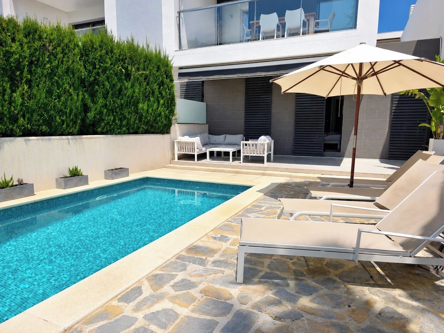 A swimming pool with lounge chairs and an umbrella, surrounded by a stone patio and a hedge.
