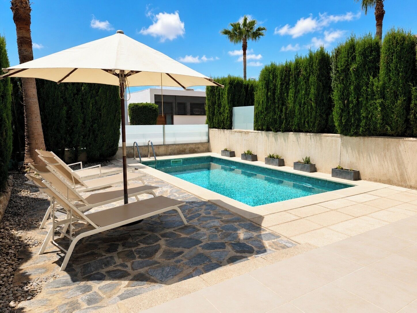 A swimming pool with lounge chairs and an umbrella, surrounded by stone tiles and greenery.