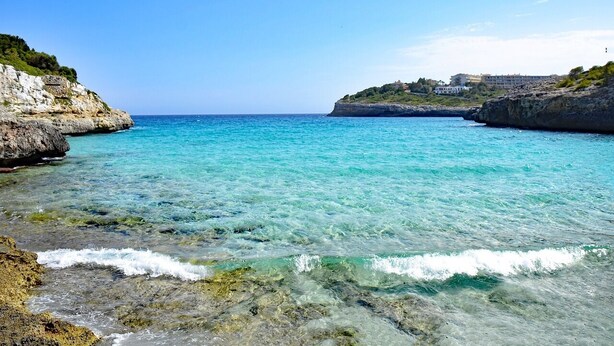 A coastal scene with clear turquoise water, rocky shores, and buildings on a hill in the background. - Casa Descansa - vacation to feel good, enjoy and relax! (Cala Mendia)