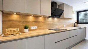A modern kitchen with a wooden countertop, built-in cabinets, a black range hood, and a window with blinds. - Casa Descansa—a vacation to feel good, enjoy yourself, and relax! (Cala Mendia)