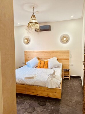 Un dormitorio con una cama de madera, ropa de cama blanca, un taburete de madera pequeño y una lámpara de techo tejida original.