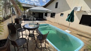 A swimming pool with a table and chairs, surrounded by a patio area with a covered seating area and umbrellas. - House in gated community with privileged location (Aparecida de Goiânia)