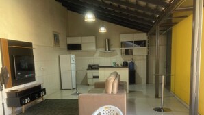 A compact kitchen with white cabinets, a microwave, and a gas stove. There is a dining area with a round table and four white plastic chairs. - House in gated community with privileged location (Aparecida de Goiânia)