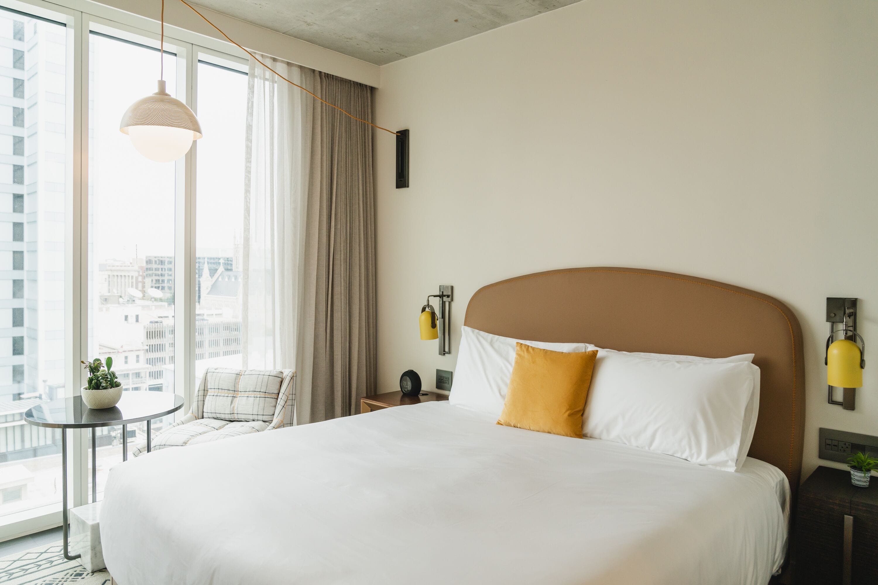 A hotel room with a large bed, a small table with a plant, a chair, and a view of the city through large windows.