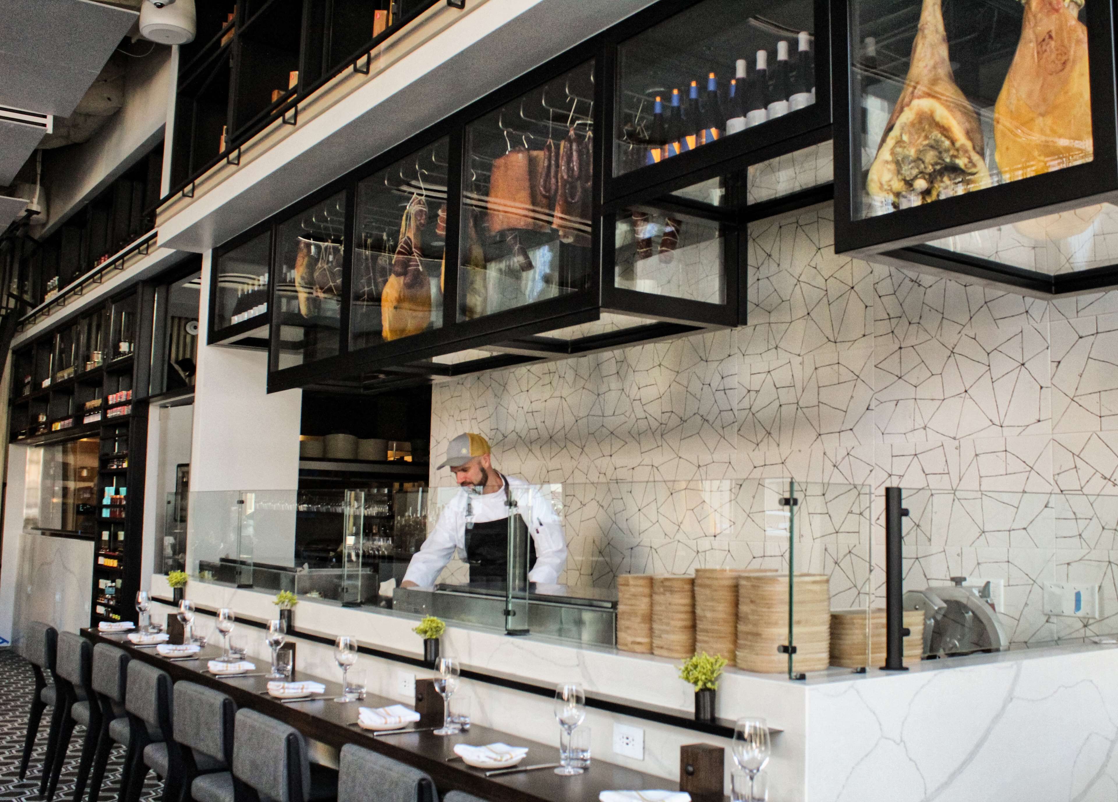 Un restaurant au décor moderne, avec un chef cuisinant, une longue table à manger entourée de chaises et un présentoir à bouteilles de vin.