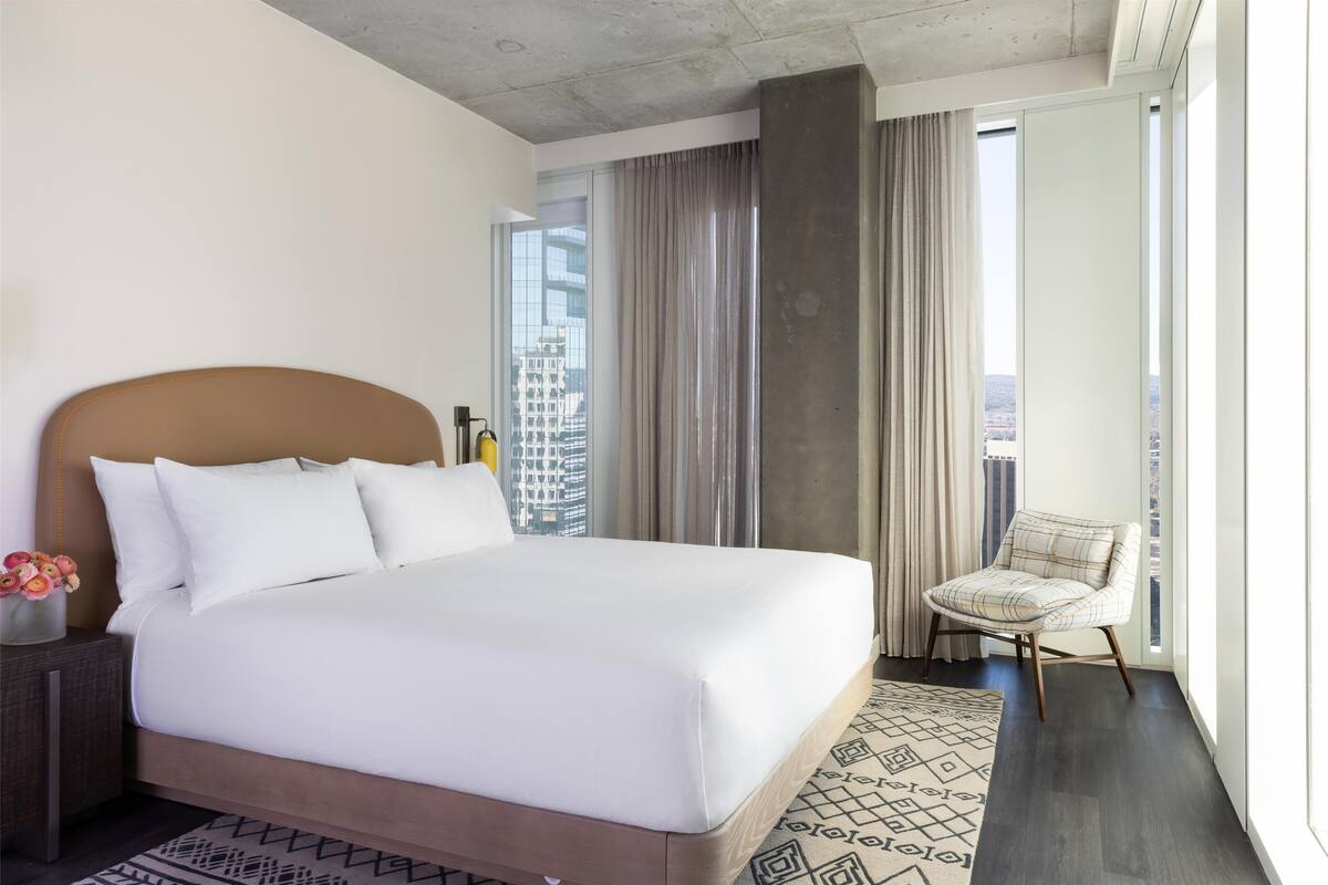A modern hotel room with a large bed, a nightstand, a chair, and a view of the city.